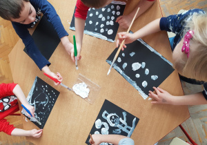 Dzieci malują zimę białą farbą na czarnym kartonie.